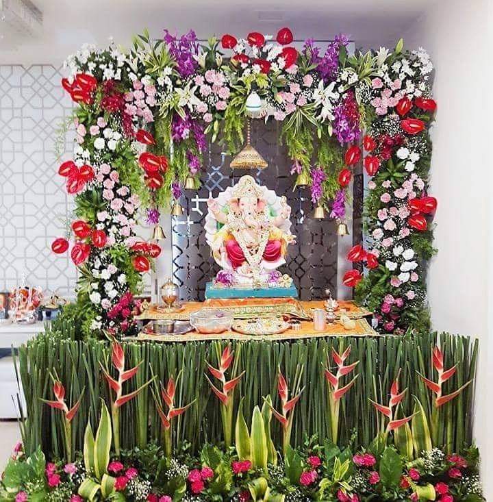 Premium Ganpati floral mandap decoration with red anthuriums, pink carnations, white lilies, orchids, heliconia, green foliage, and brass bells for festive celebration.