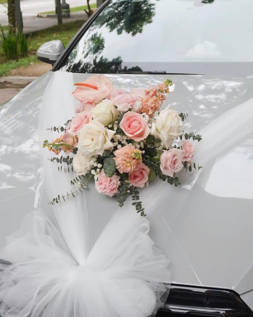 Stylish wedding car decoration featuring pastel pink roses, white roses, peach carnations, pink anthurium, baby’s breath, and eucalyptus arranged on the bonnet with white tulle fabric.