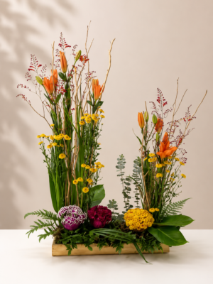 Modern floral arrangement featuring orange lilies, yellow button chrysanthemums, ornamental kale, decorative branches, eucalyptus, and fresh greenery in a wooden planter.