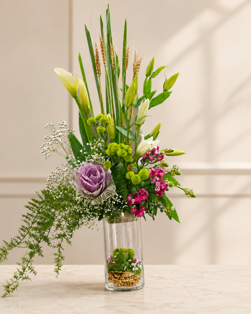 A fresh flower arrangement featuring white lilies, calla lilies, ornamental cabbage, green button chrysanthemums, pink dianthus flowers, baby's breath, wheat stems, and lush greenery in a tall clear glass vase.