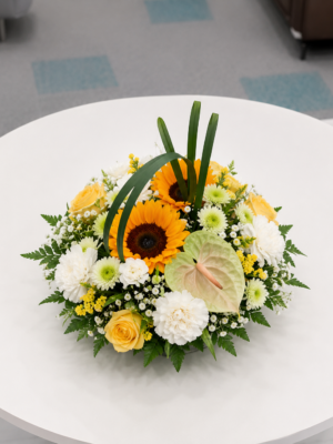 Sunflower floral table centerpiece with yellow roses, white carnations, chrysanthemums, anthurium, and fresh greenery arranged in a low glass bowl on a round table