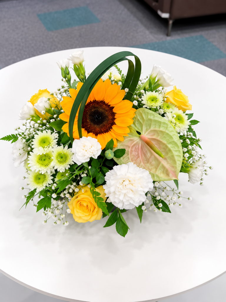Sunflower floral table centerpiece with yellow roses, white carnations, chrysanthemums, anthurium, and fresh greenery arranged low on a round table