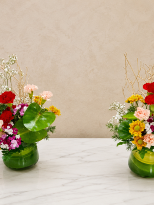 Modern mixed flower table arrangement featuring carnations, chrysanthemums, dianthus, baby's breath, anthurium, seasonal flowers, decorative branches, and greenery in a green glass vase.