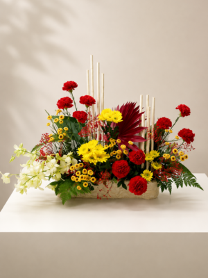 Modern floral centrepiece with red carnations, yellow daisies, white orchids, decorative reeds, and lush greenery in a rectangular container.