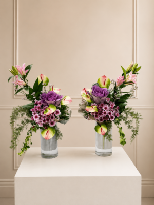 Fresh floral arrangement featuring pink lilies, pink anthuriums, ornamental cabbage, purple chrysanthemums, cascading greenery, and seasonal foliage arranged in a tall clear glass vase.