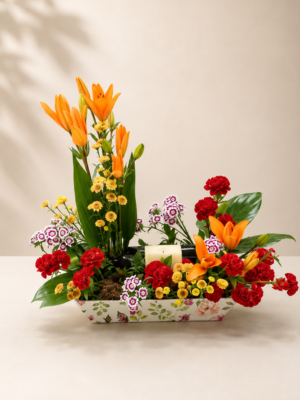 Premium floral centerpiece featuring orange lilies, red carnations, yellow button chrysanthemums, Sweet William flowers, and fresh greenery in a decorative ceramic tray.
