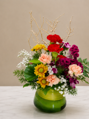 Modern mixed flower table arrangement featuring carnations, chrysanthemums, baby's breath, seasonal flowers, decorative branches, and greenery in a green glass vase.