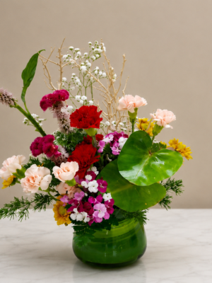 Modern mixed table flower arrangement featuring red and peach carnations, chrysanthemums, baby's breath, seasonal flowers, decorative branches, and greenery in a green glass vase.